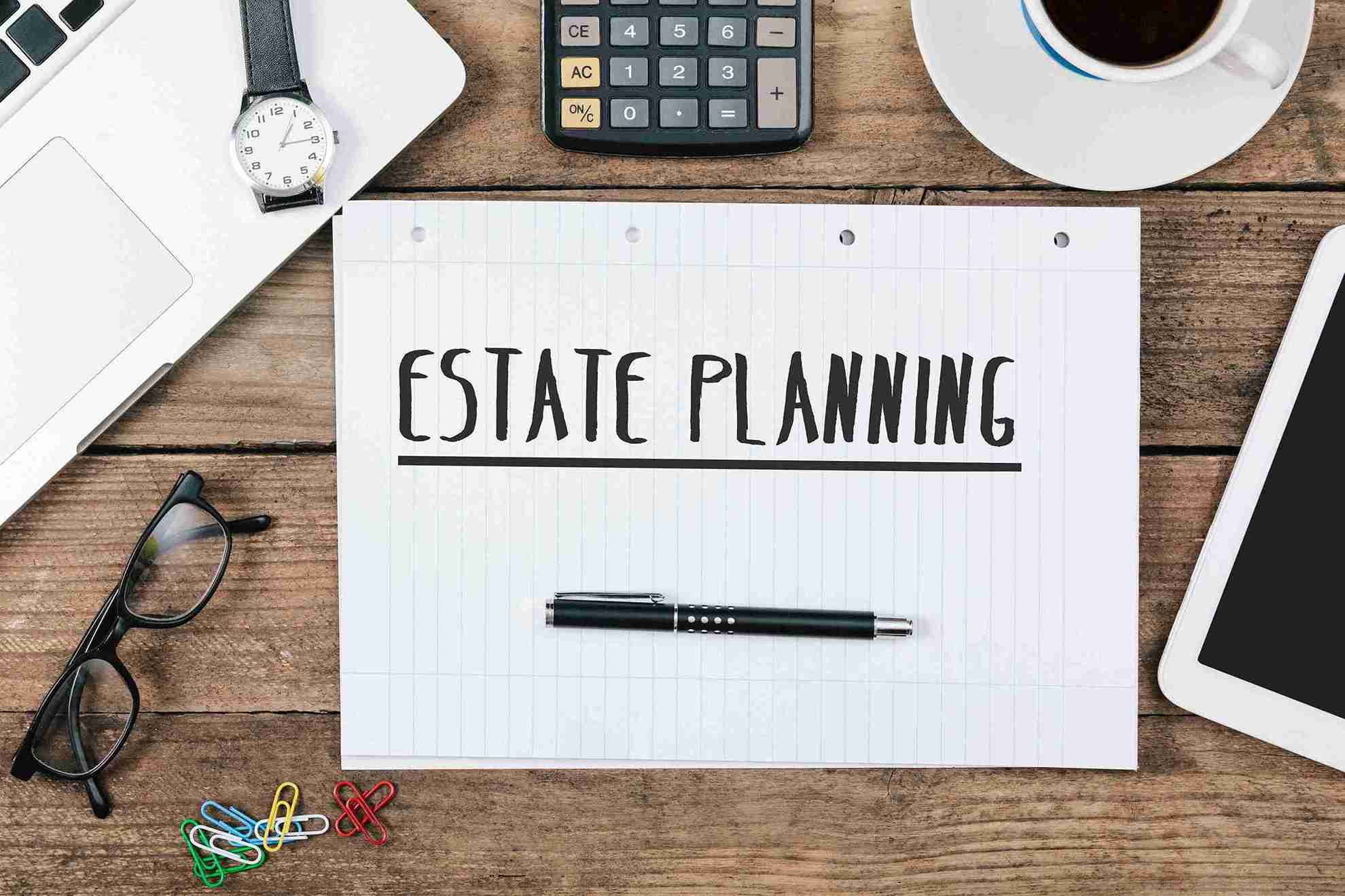 A notepad on a wooden desk displays "Estate Planning" with a pen on top. Surrounding items include a laptop, watch, calculator, glasses, and a coffee cup.