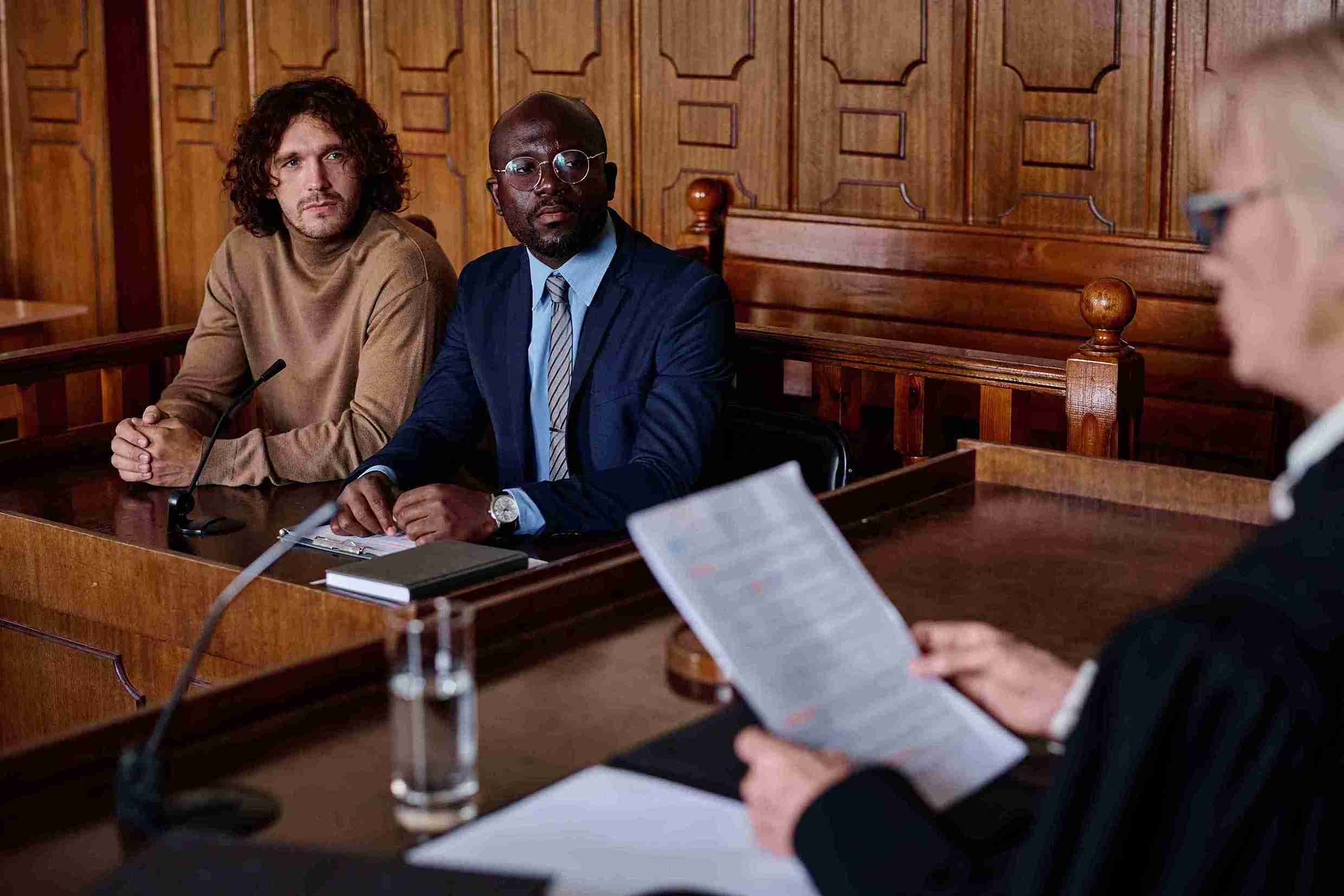 Two men seated in a courtroom, attentively listening to a judge presiding over a legal matter.