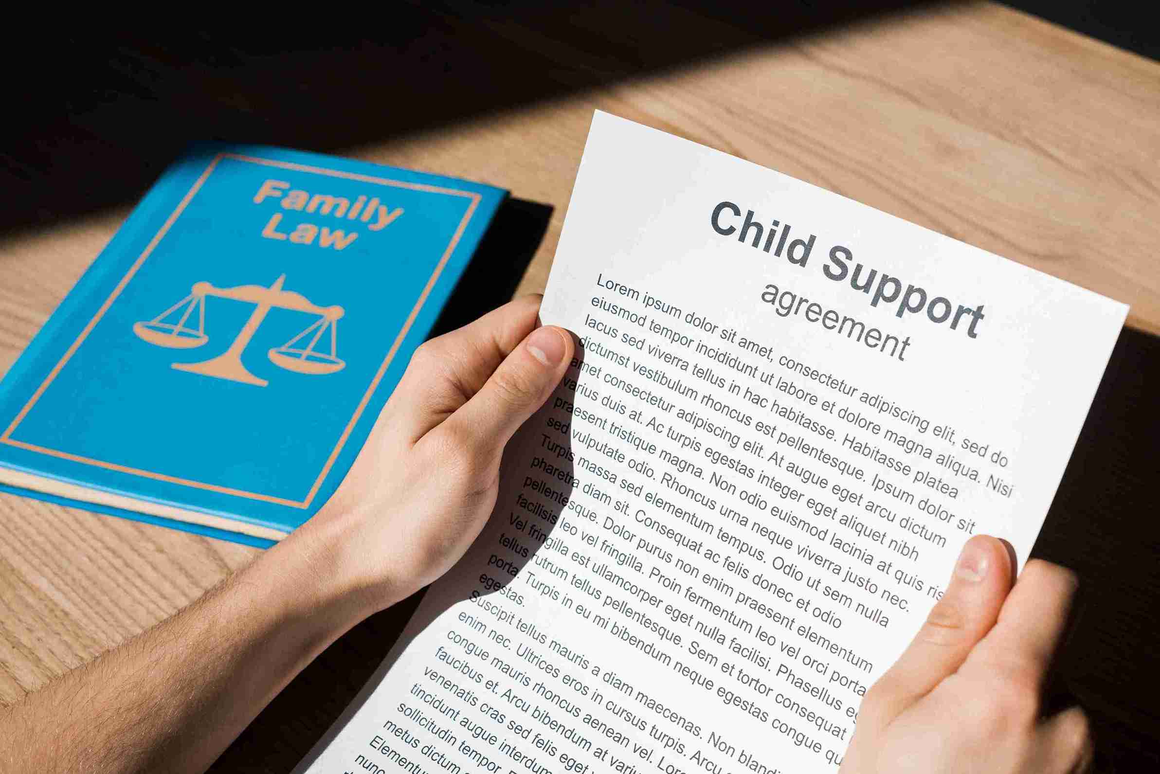 Hands holding a "Child Support Agreement" document over a table. Next to it is a blue book titled "Family Law" with scales symbol, suggesting a legal context.