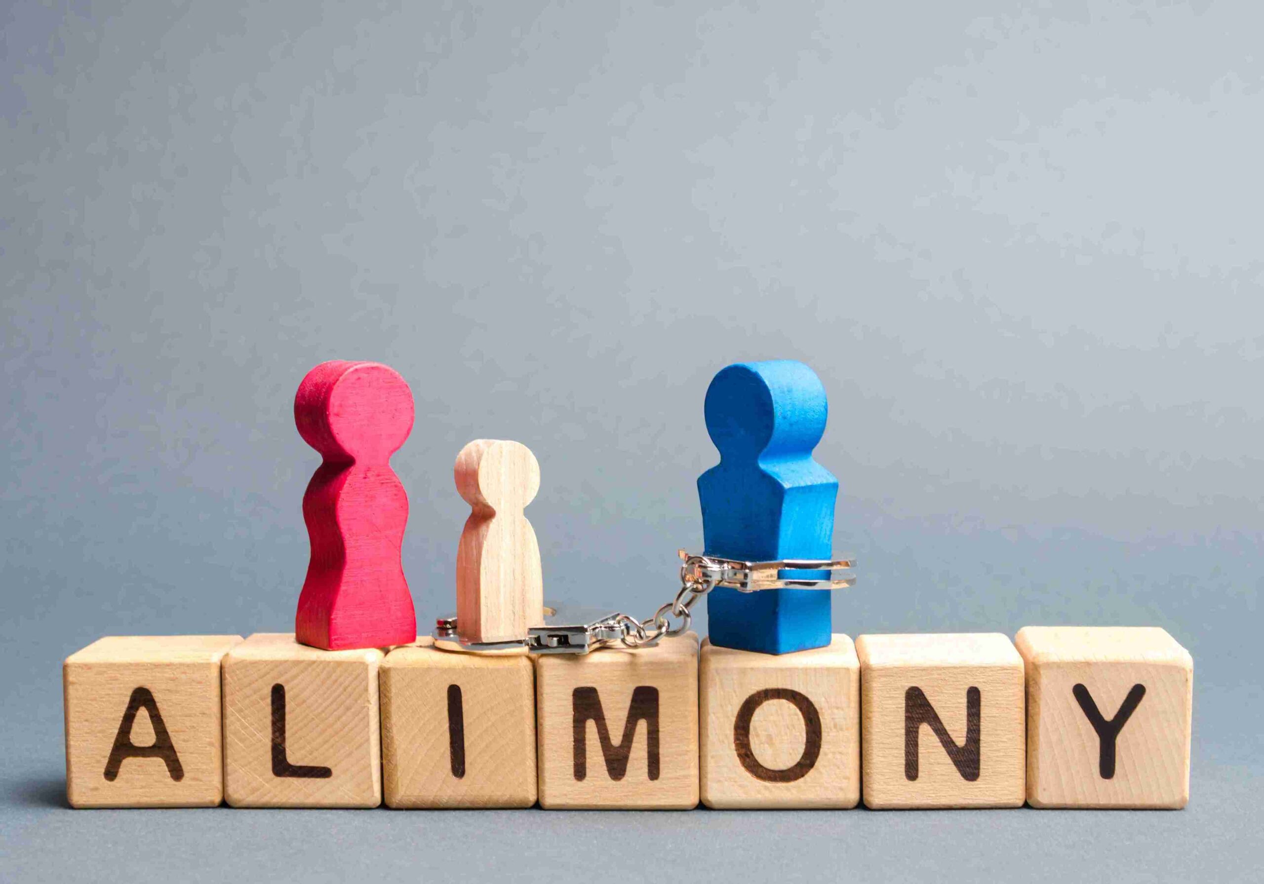 Wooden figures representing a family stand on blocks spelling "ALIMONY." Two larger figures, red and blue, are connected by handcuffs, symbolizing financial obligation.