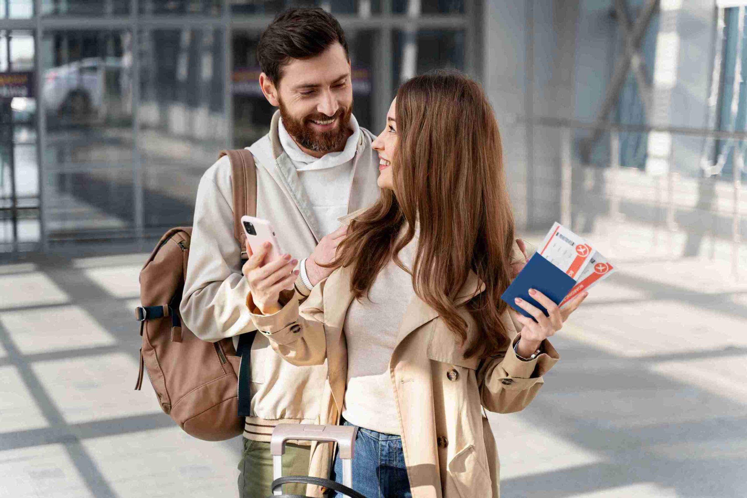 A smiling couple at an airport, holding passports and tickets. The woman wears a beige trench coat; the man has a backpack. They appear excited and happy.