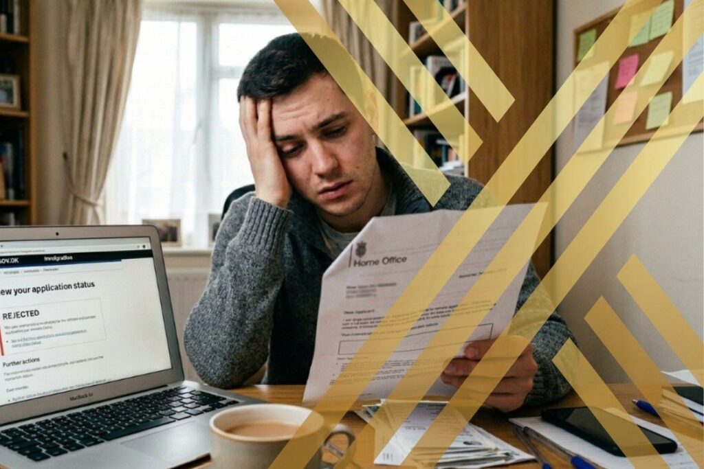 Frustrated applicant sitting at a desk in a home office holding an indefinite leave to remain refusal letter, with laptop open showing UK immigration website on the screen.