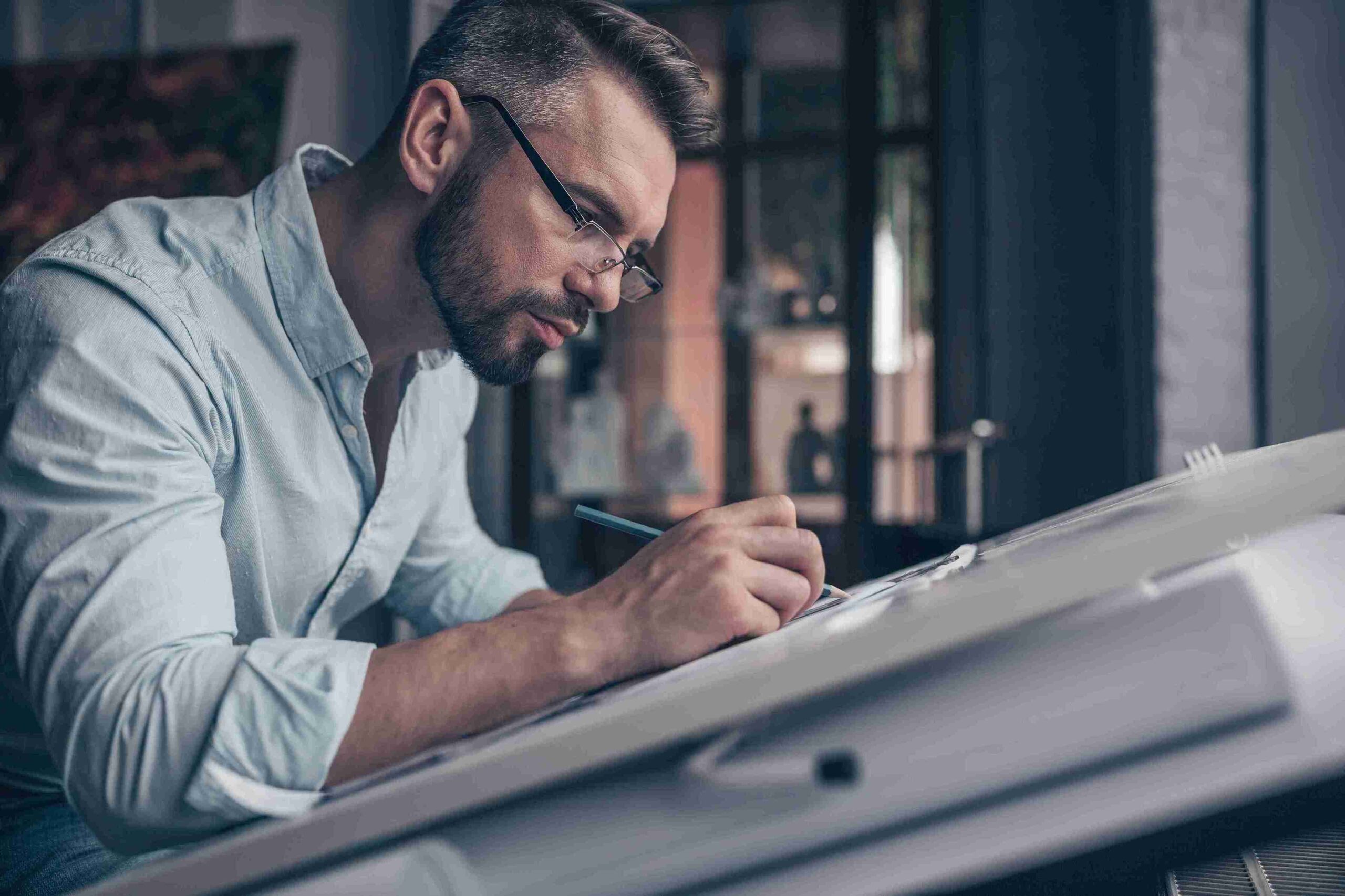 Man in glasses and blue shirt focusing intently while drawing on a drafting table in a dimly lit room, conveying creativity and concentration.
