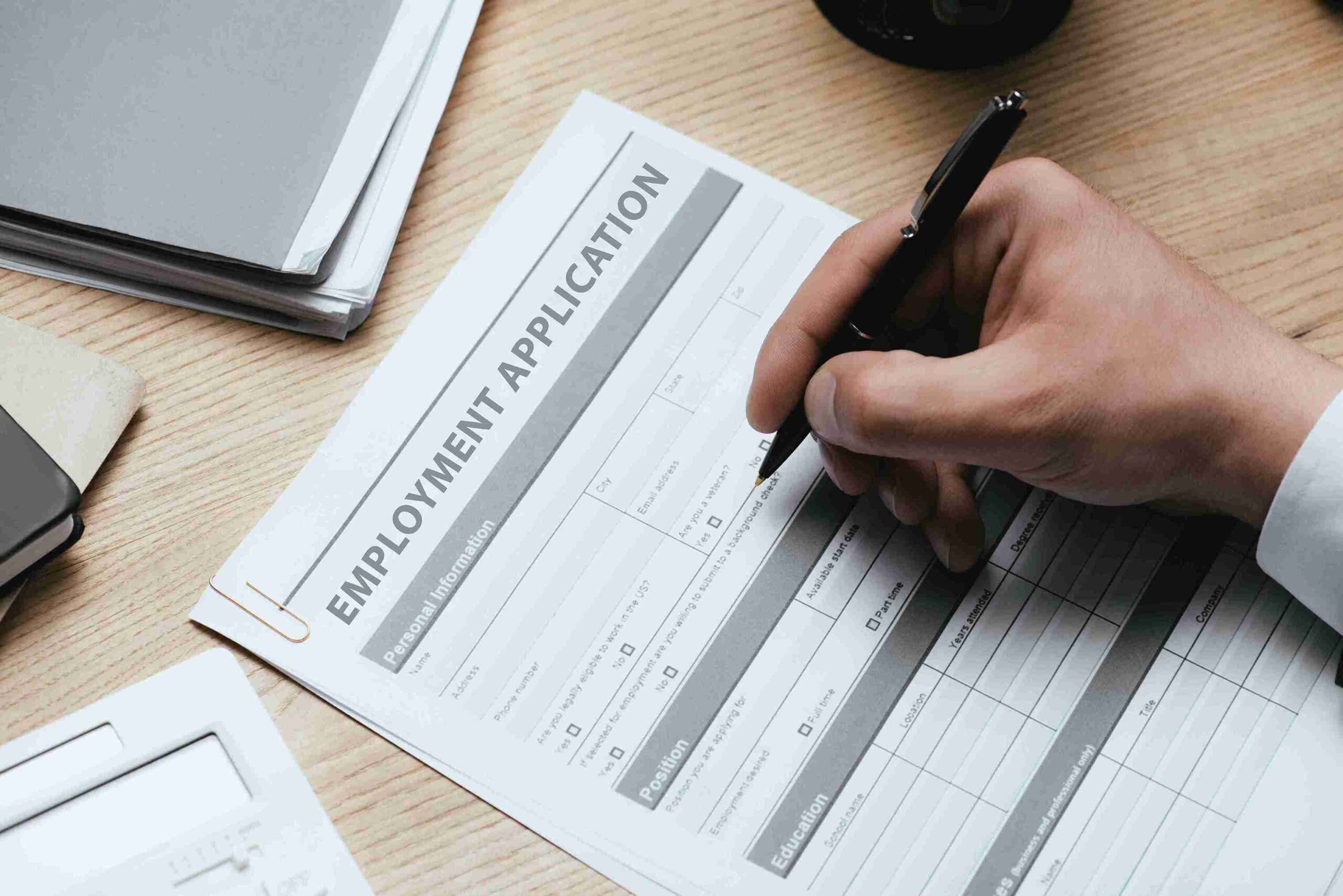 A hand holding a pen fills out an employment application form on a wooden desk.