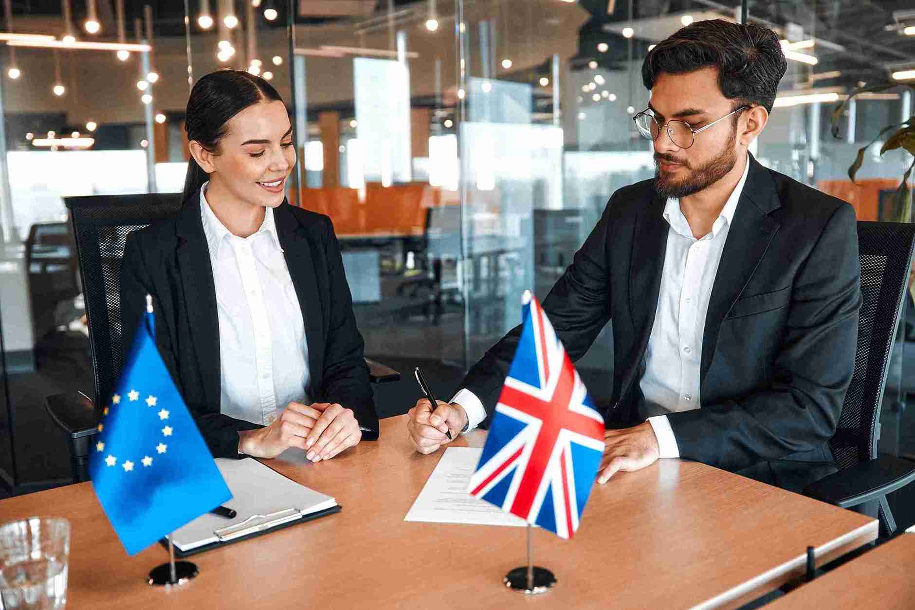 Two professionals in suits sit at a table with EU and UK flags.