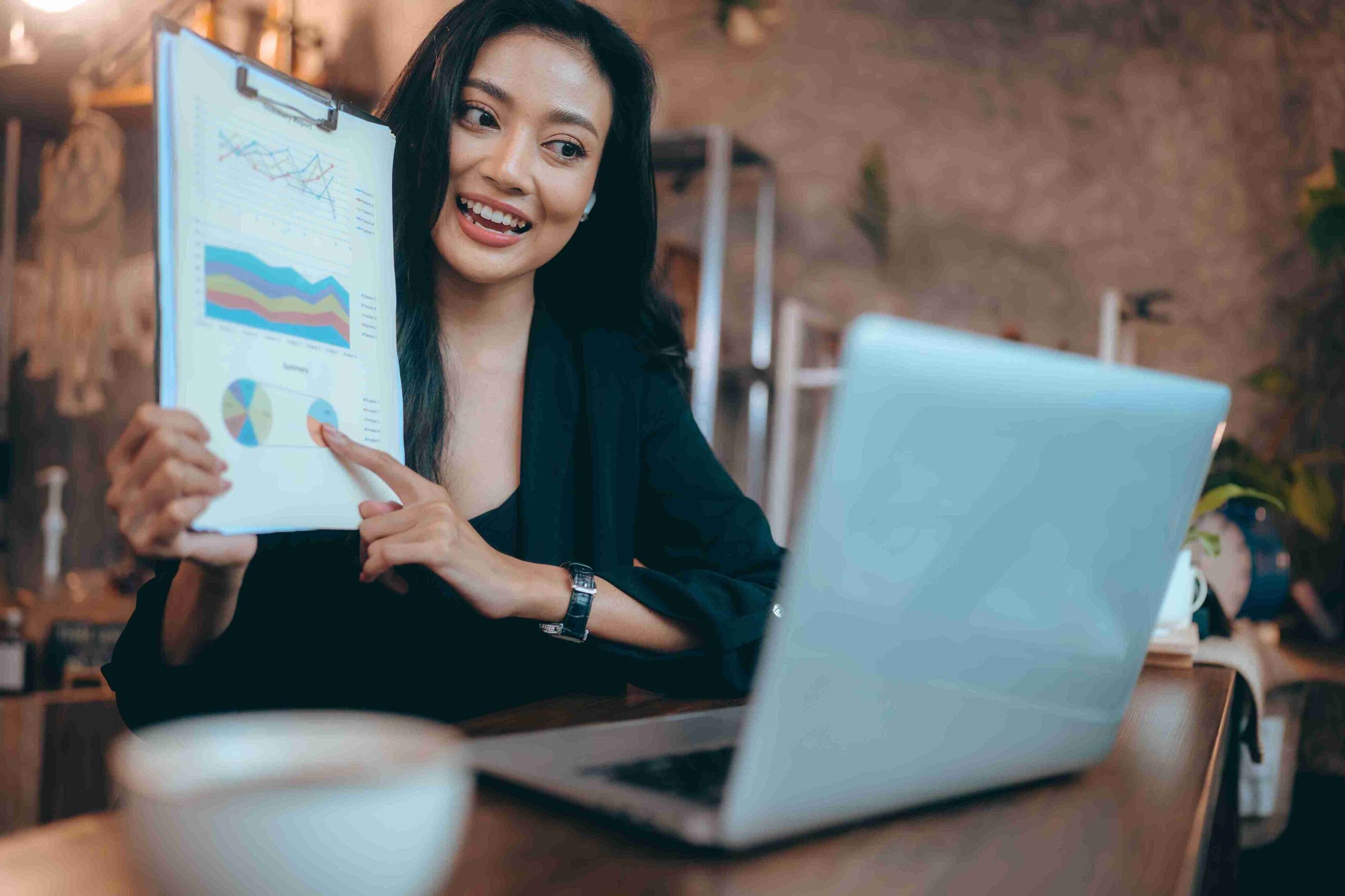 A smiling woman holds a clipboard while looking at a laptop, exuding confidence and engagement in her work.