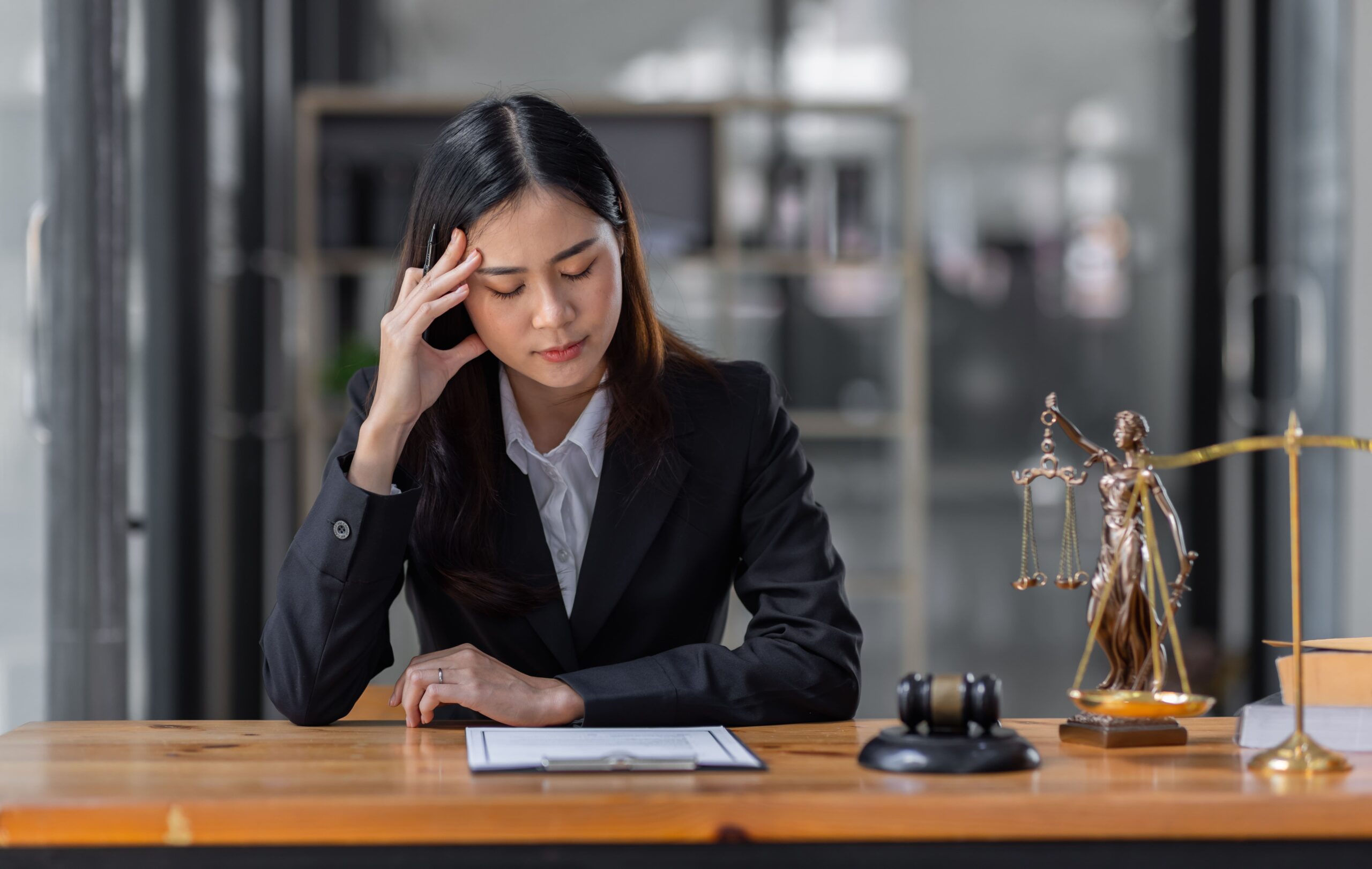 A woman in a black suit sits at a desk, looking stressed. She holds her head while reading a document. A judge's gavel and scales of justice are visible.