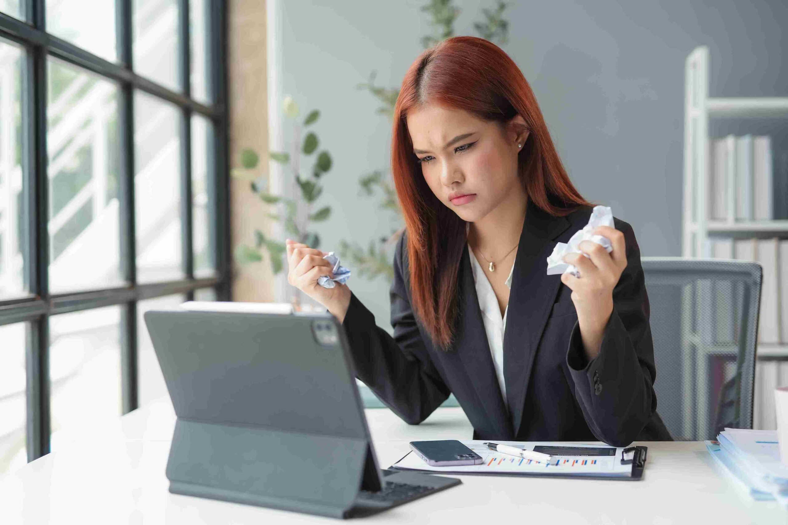 A frustrated woman in a suit sits at a desk, crumpling paper, and staring at a laptop.