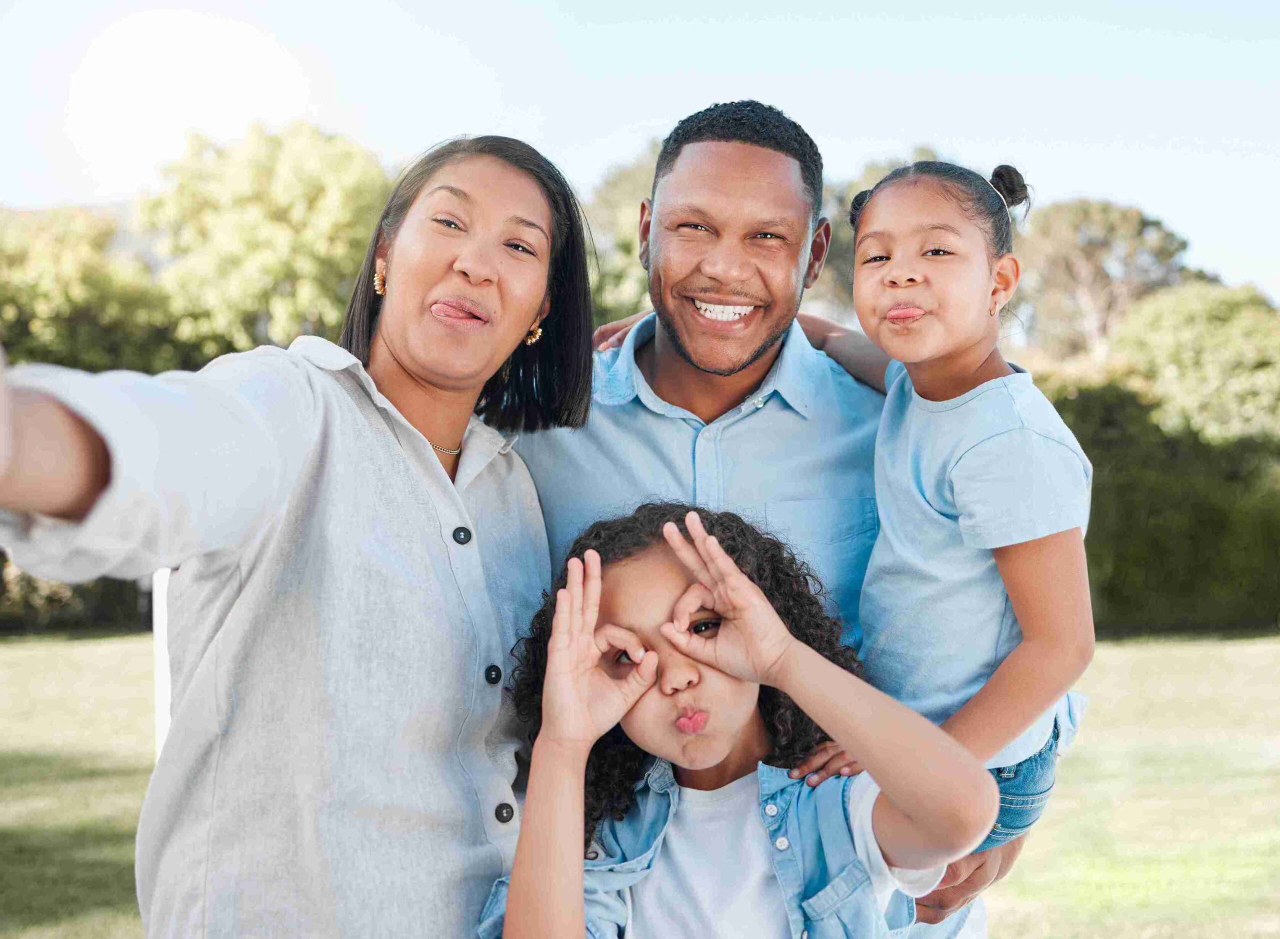 A family of four poses for a selfie outdoors, smiling and making playful faces with greenery in the background.
