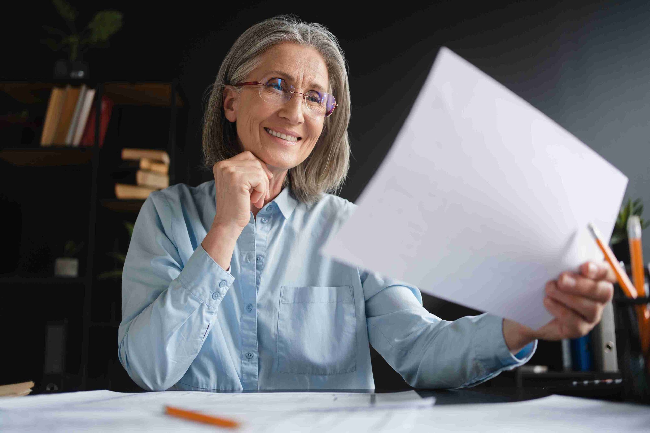A woman wearing glasses and a blue shirt holds a piece of paper, looking focused and engaged in her task.
