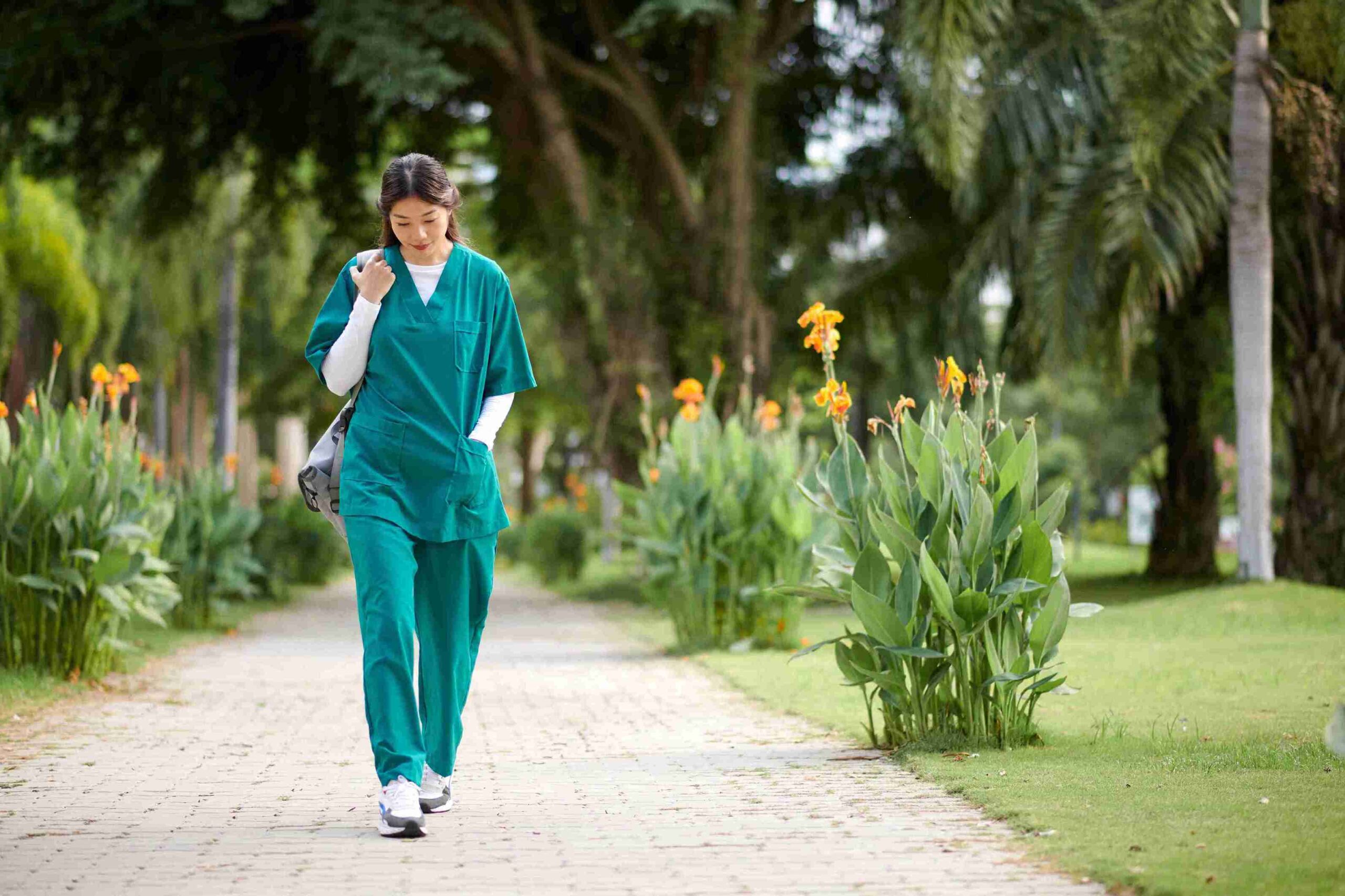 A woman in a green scrub suit walks confidently down a path surrounded by greenery.