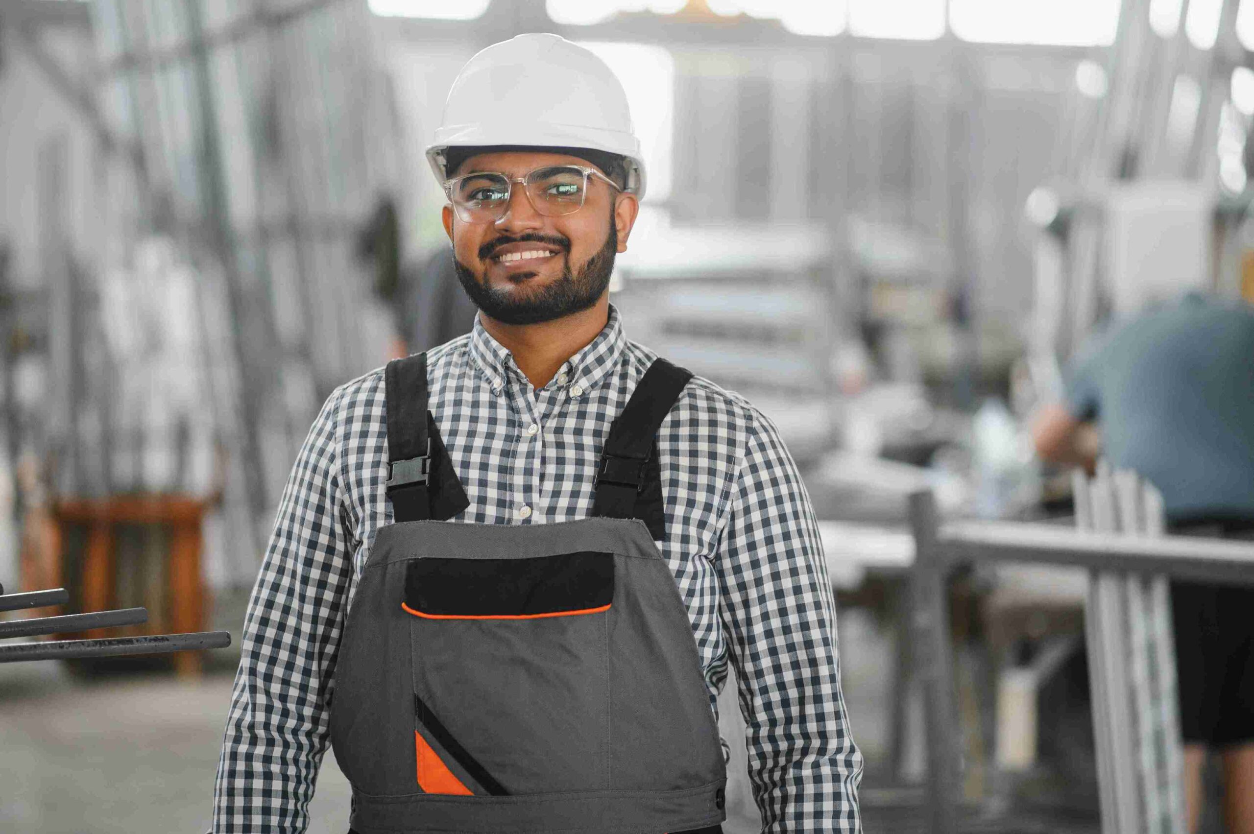 Smiling man in a hard hat and glasses stands in an industrial setting. He wears a checkered shirt and gray overalls, conveying a sense of confidence.