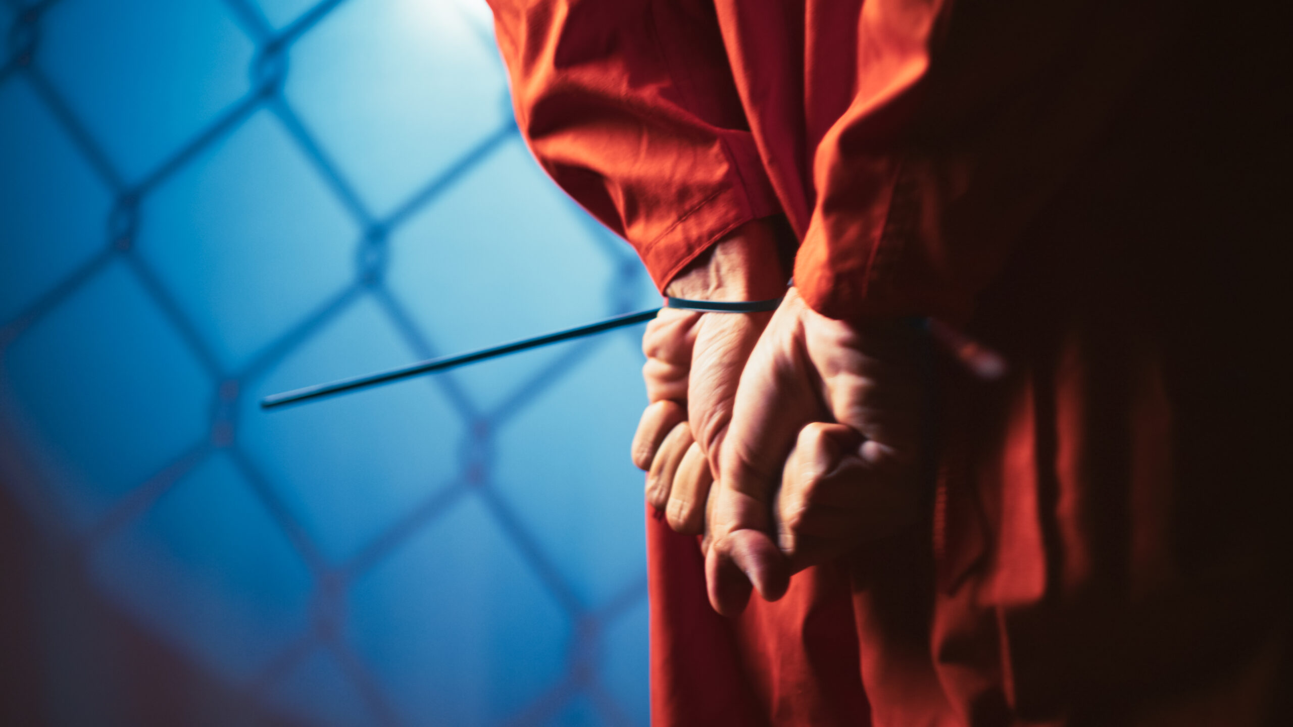 A person in an orange prison uniform stands holding a stick, looking determined against a neutral background.