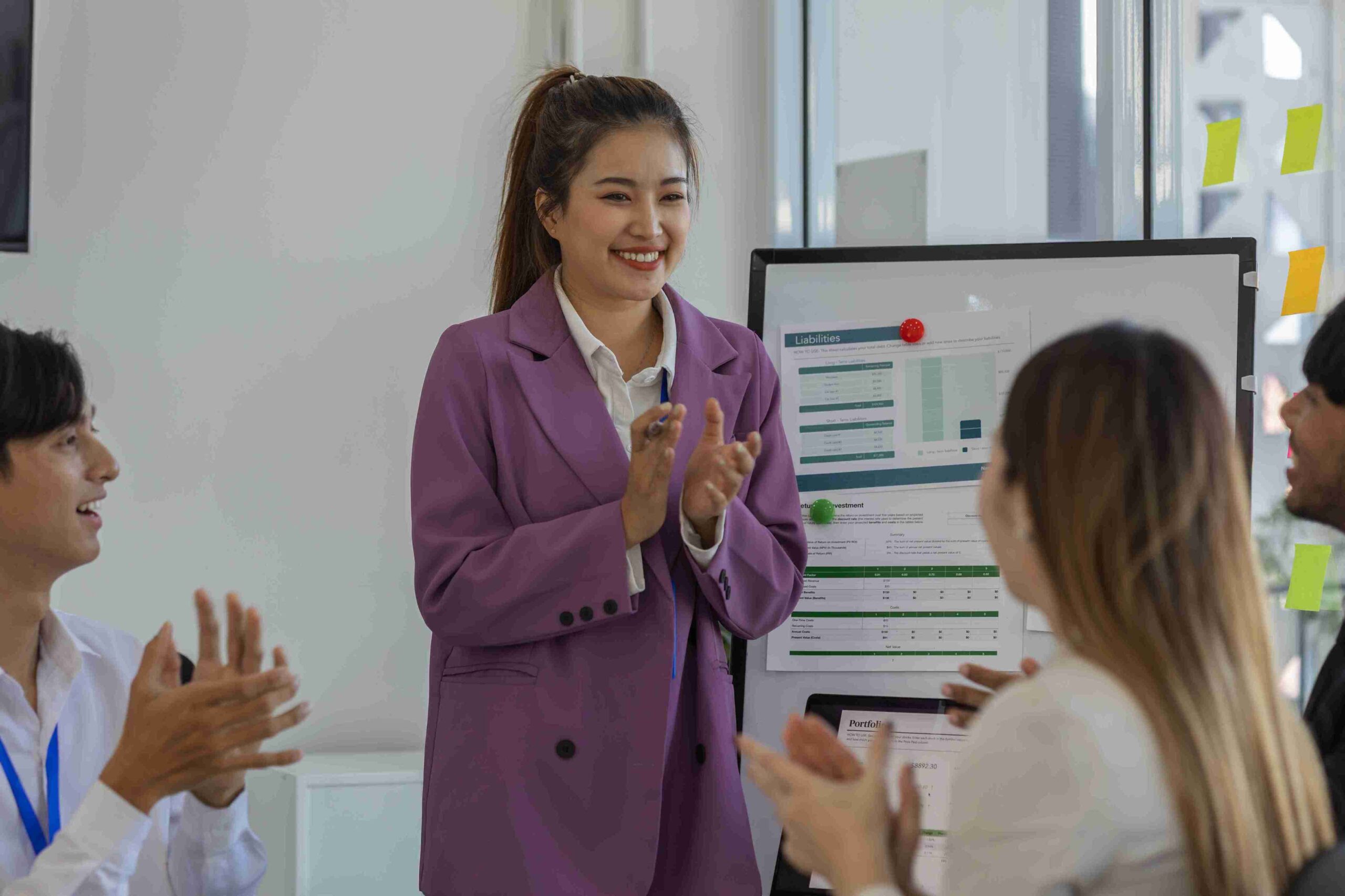 A smiling woman in a business suit claps enthusiastically, celebrating her colleagues' achievements in a professional setting