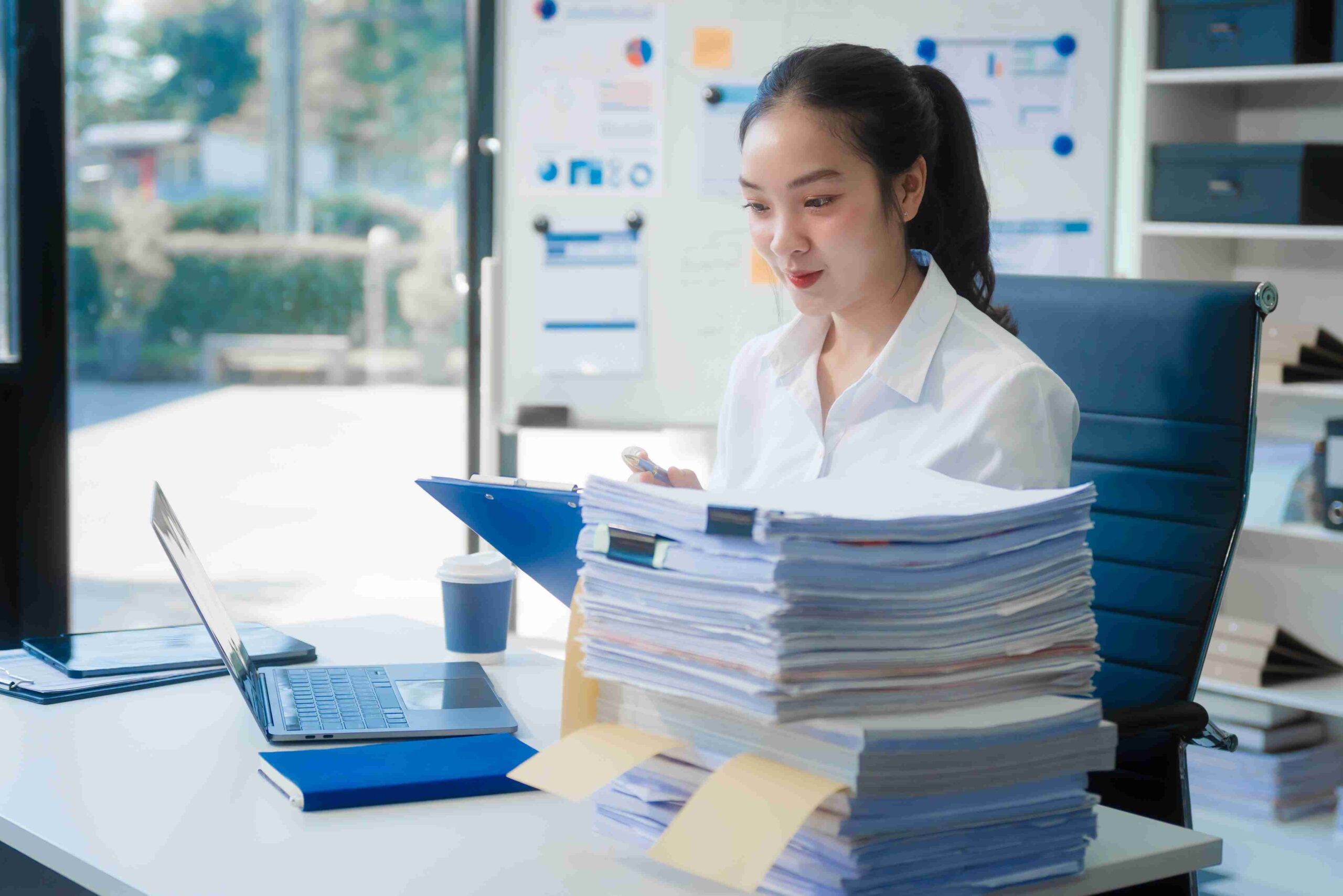 A woman sits at a desk, focused on a stack of papers in front of her, surrounded by a tidy workspace.