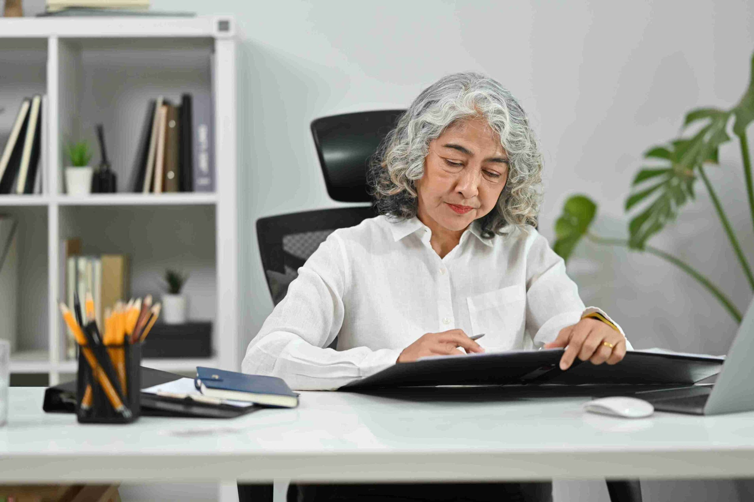 An older woman with gray hair sits at a desk, focused on a digital tablet. She wears a white shirt, surrounded by office supplies and plants.