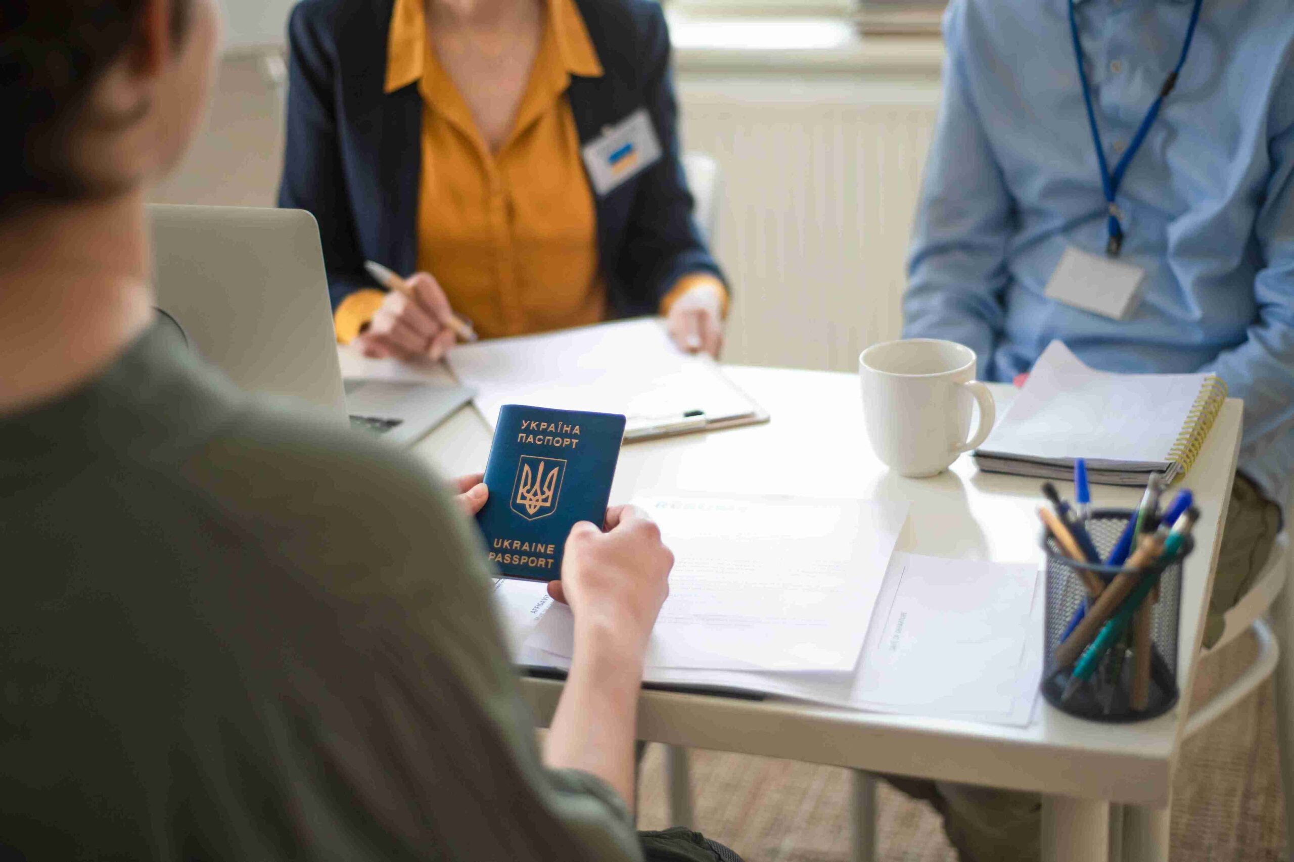 A person holds a Ukrainian passport at a table with paperwork and a laptop.