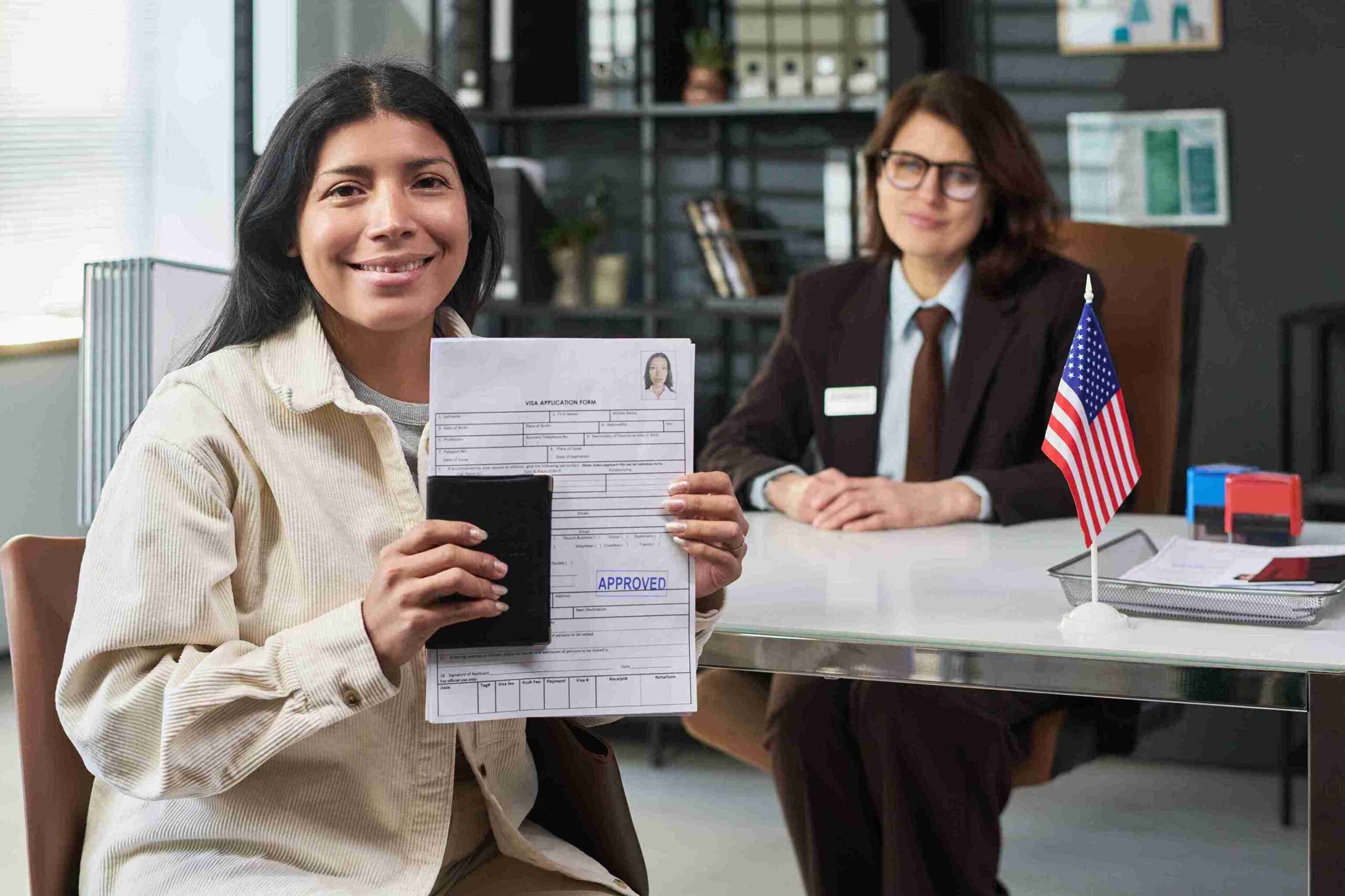 A woman smiling holds an "Approved" passport and visa document. She's seated in an office with a desk featuring a small American flag, conveying success and relief.