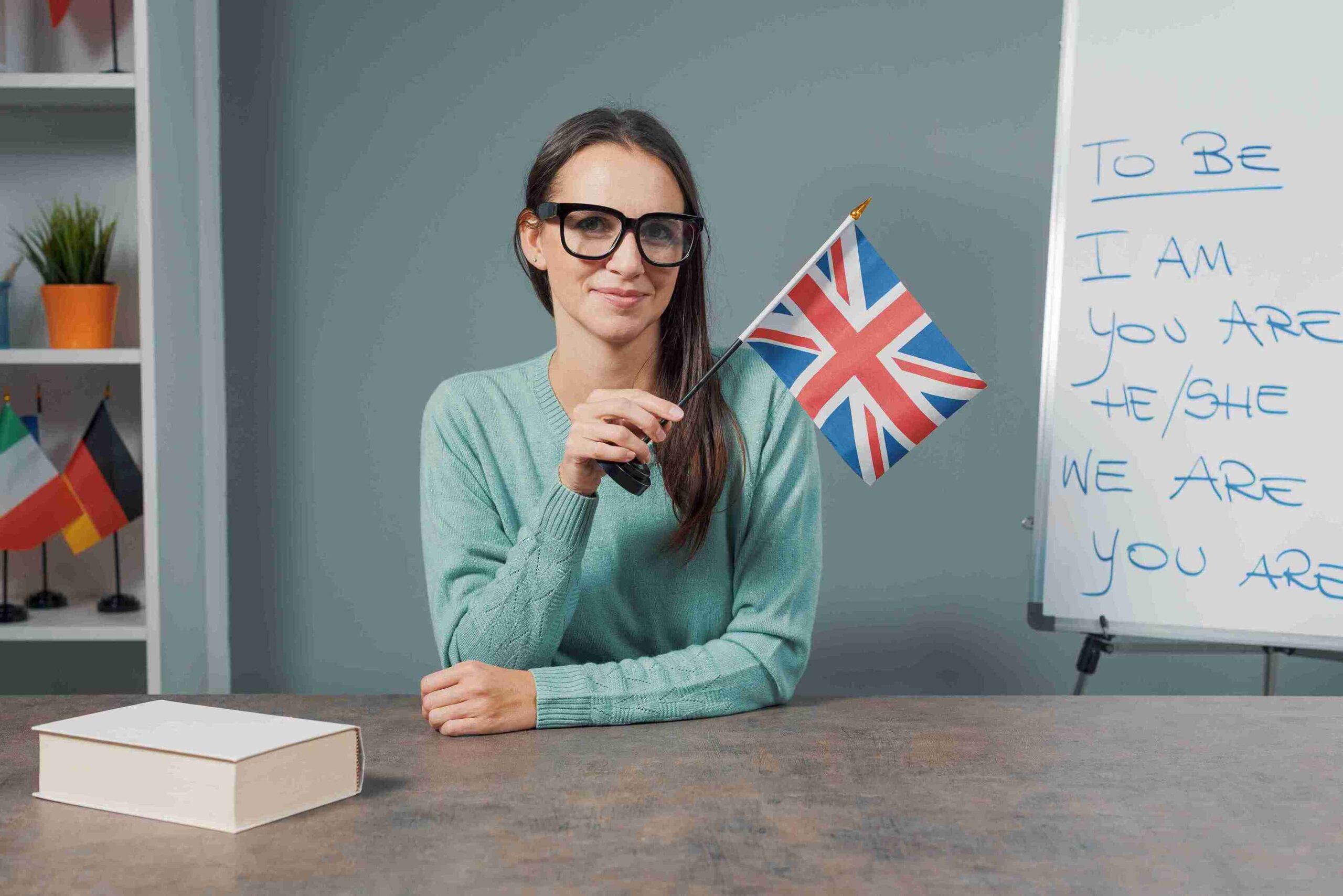 A woman with glasses stands in front of a whiteboard, holding a British flag, smiling and engaging with the audience.