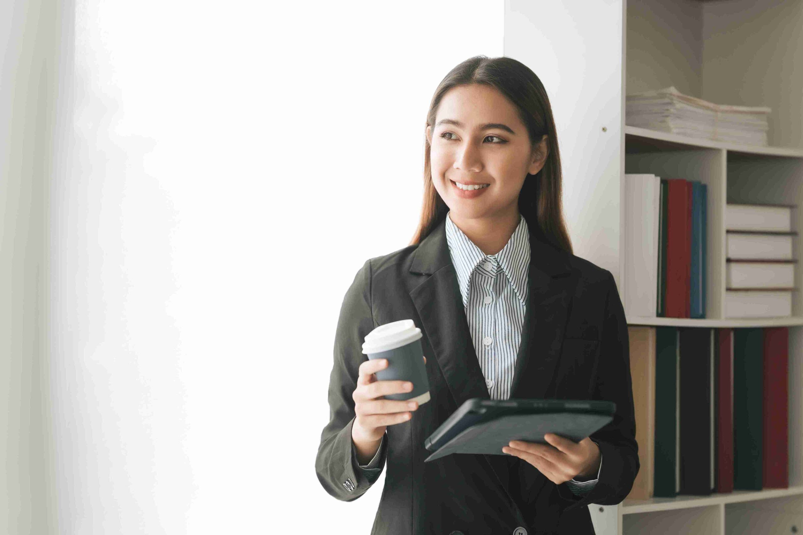 A woman in a business suit holds a cup in one hand and a tablet in the other, looking confidently at the camera.